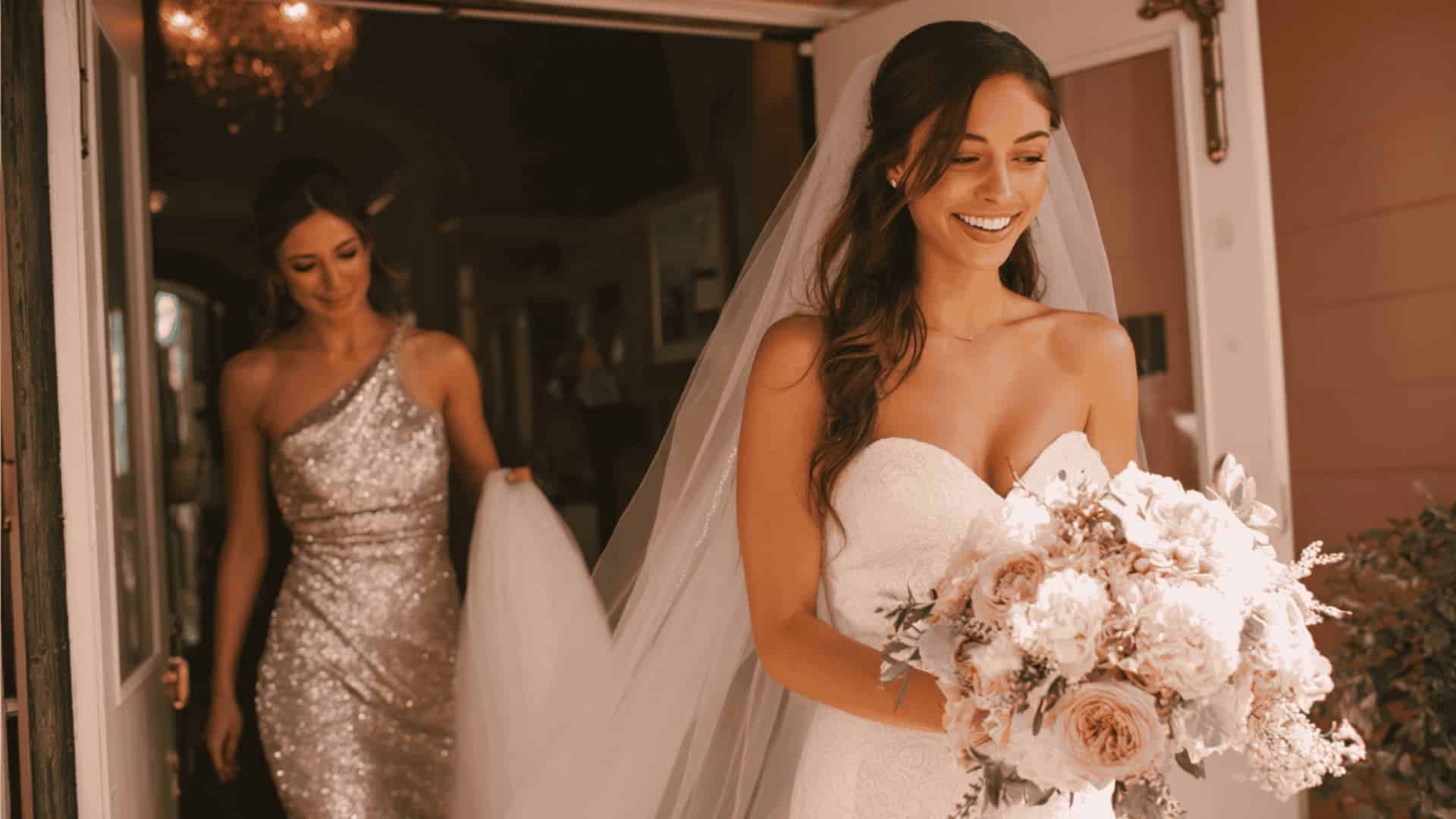 A bride in a strapless white dress with a bouquet, followed by a bridesmaid in a silver dress.