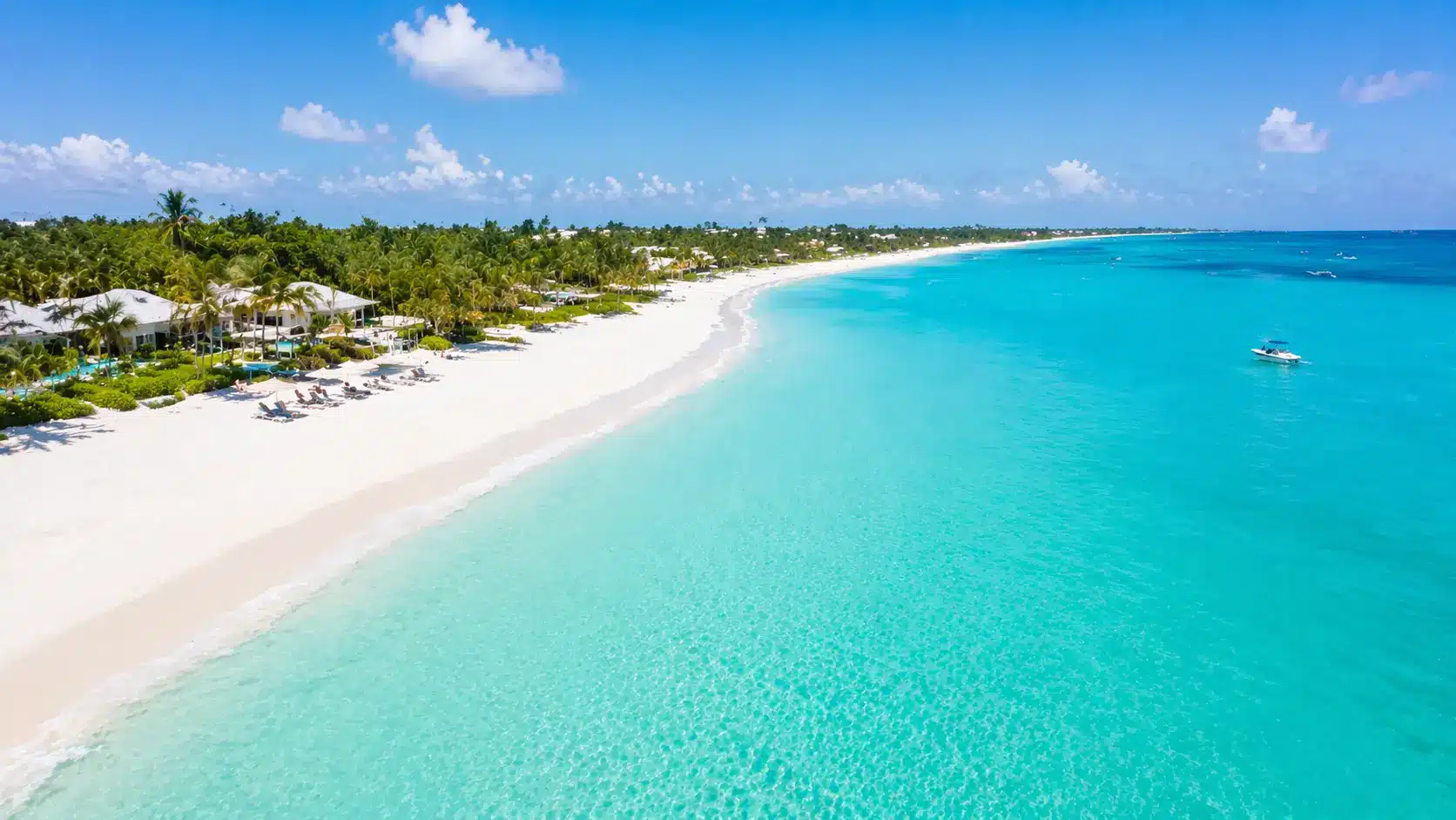 A serene aerial view of Grace Bay's pristine white sandy beach, turquoise waters, and a resort nestled among lush palm trees under a bright blue sky.
