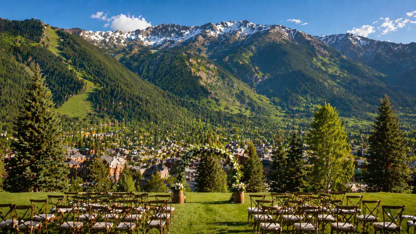 Aspen wedding scene with rows of ceremony chairs, a floral arch, and snow-capped mountains towering over a lush green valley.