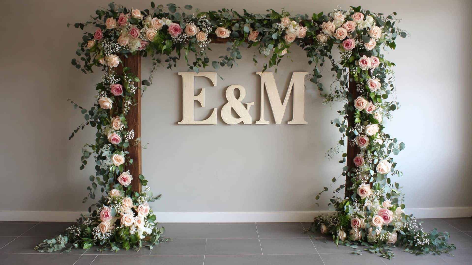 Blush rose wedding arch with greenery framing cream E and M initials on wall art