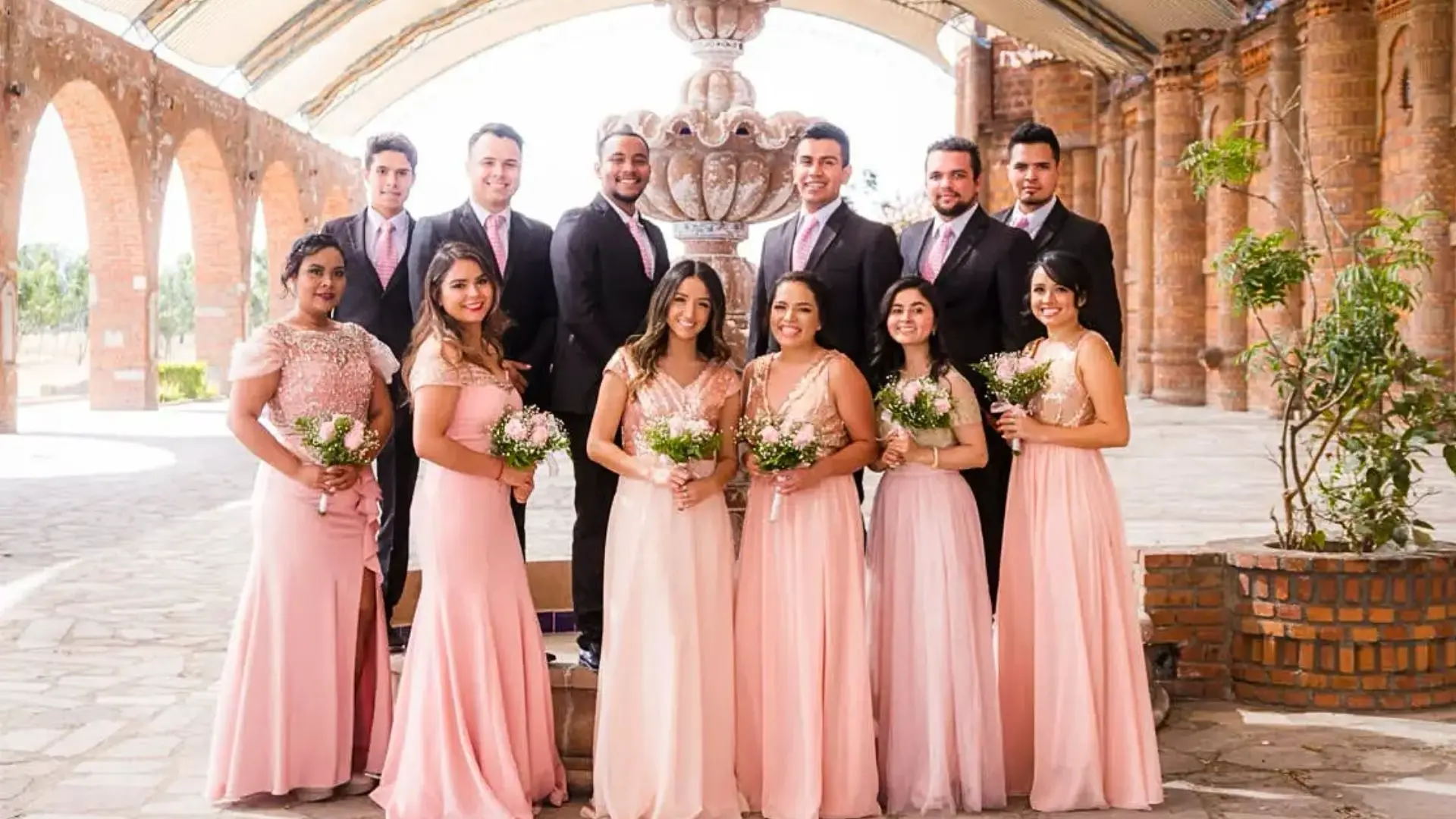 Bridal party in coordinated pink dresses and dark suits posing in front of a decorative fountain under an arched brick pavilion