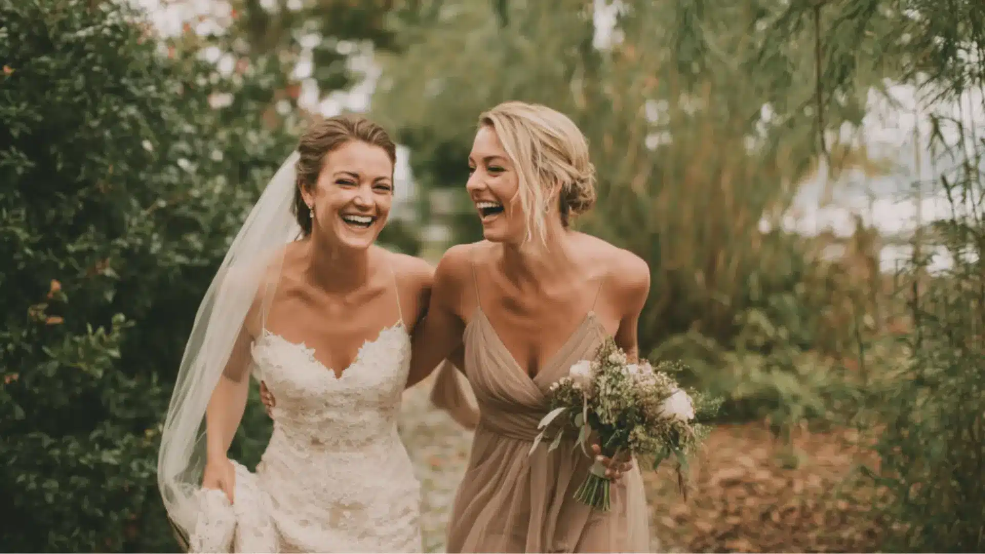 Bride and maid of honor smiling outdoors holding bouquets, with maid of honor wearing a dusty blue dress.