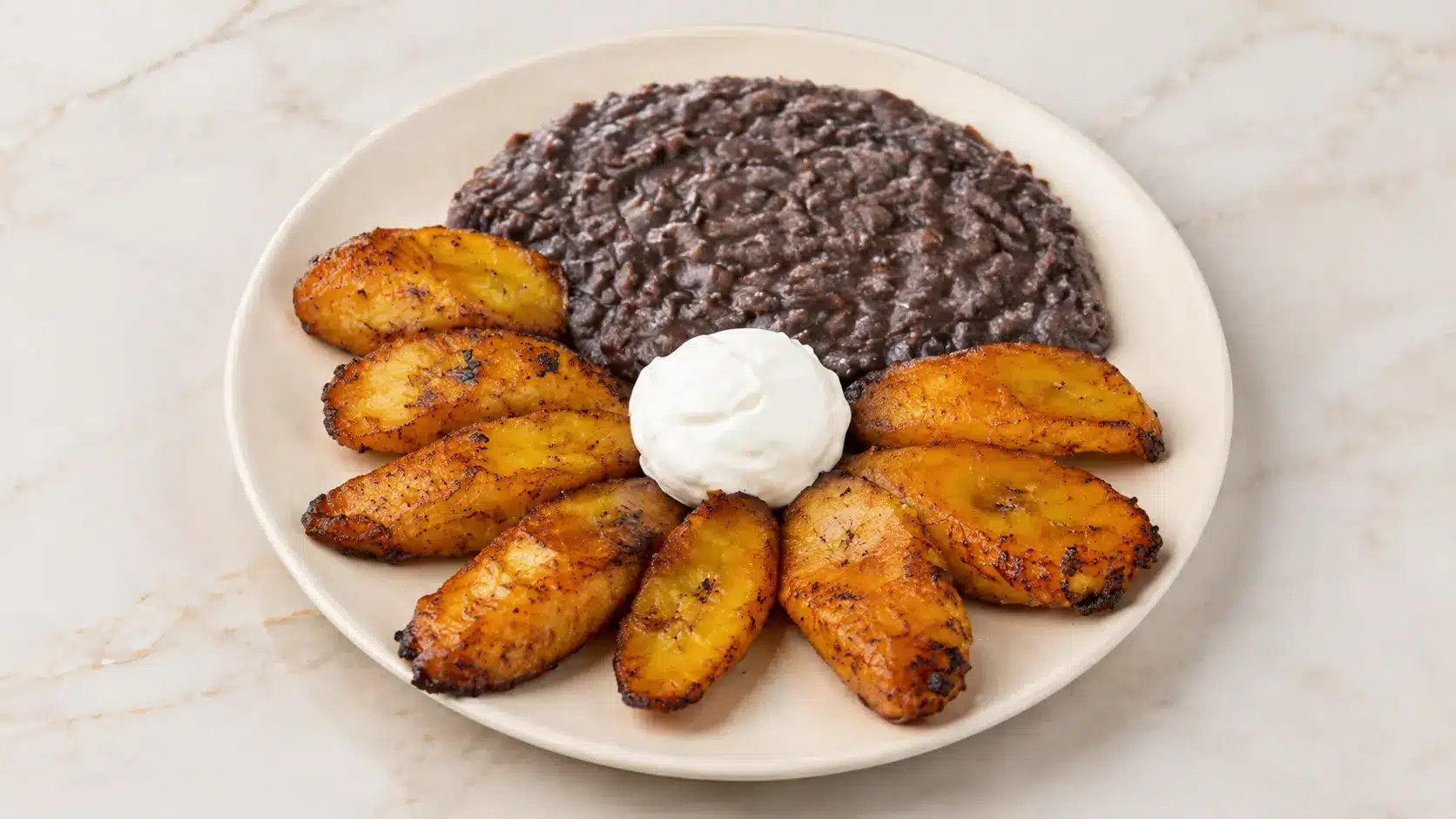 Fried plantains, refried beans, and sour cream on beige plate.