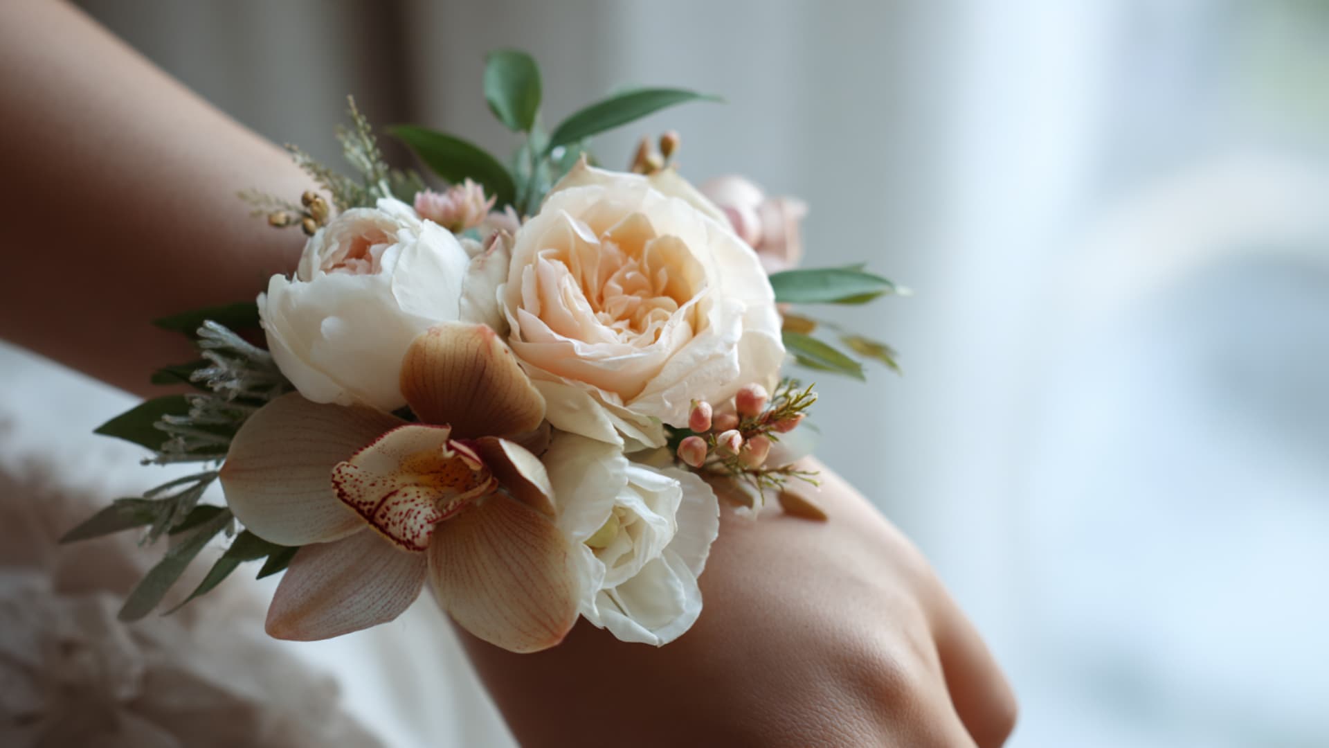Close-up of a wrist corsage with peach roses, white peonies, orchids, and greenery.