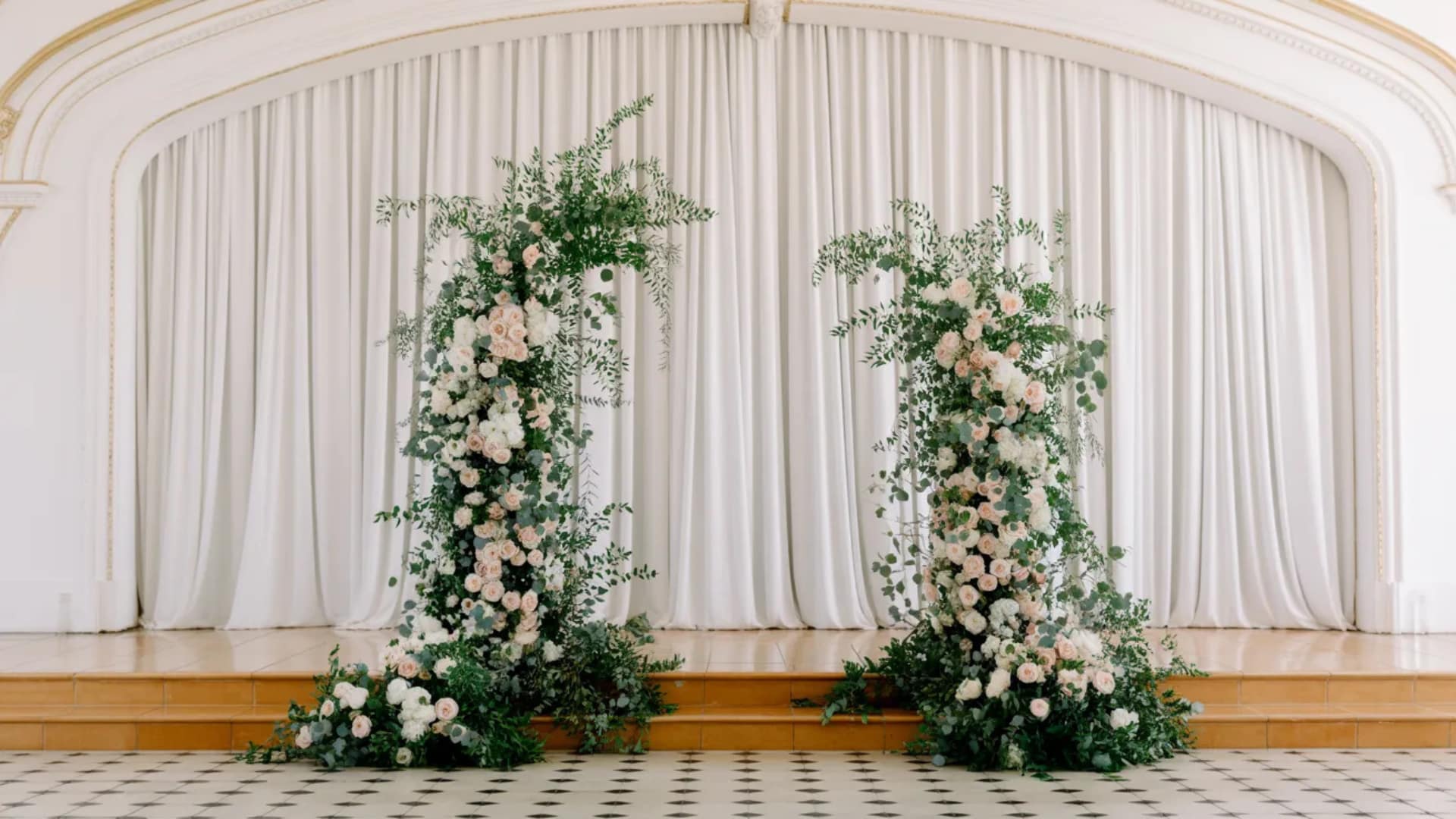 Elegant floral pillar wedding arch set before white drapes in a bright hall.