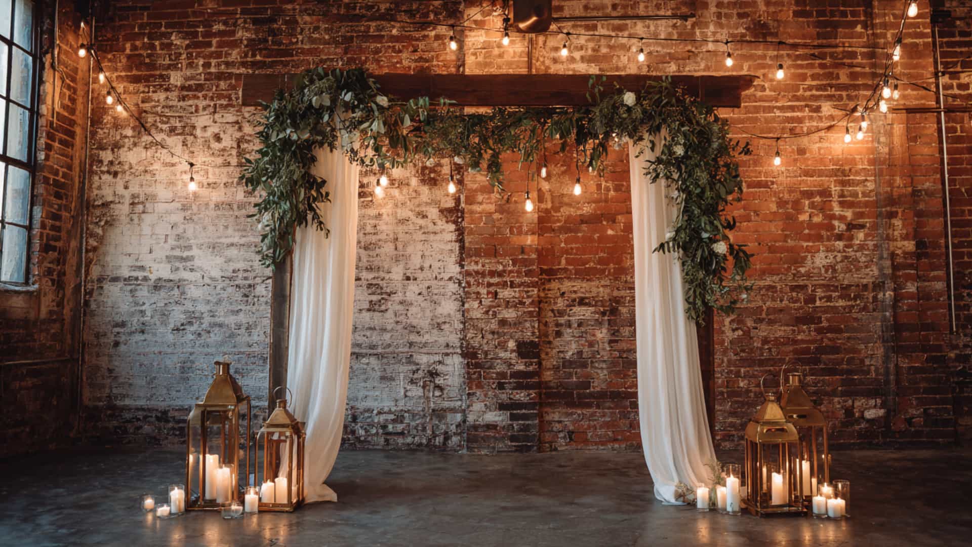 Rustic indoor wedding arch with white drapes, greenery, lanterns, and candles