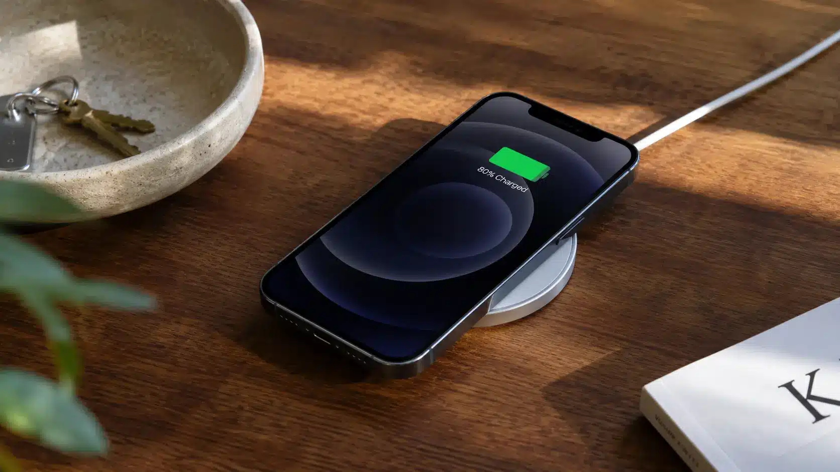 Smartphone wirelessly charging on a wooden table next to a key bowl and a book.