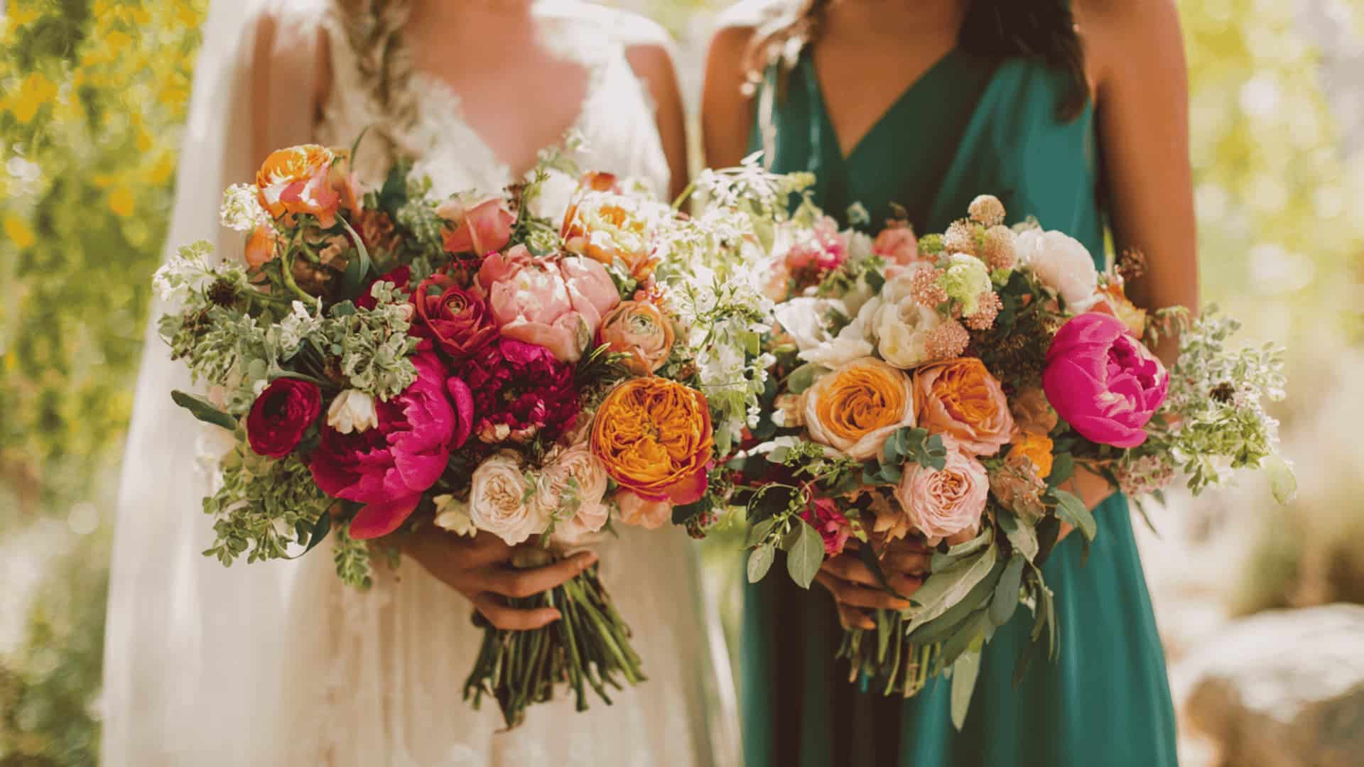 Two women in wedding attire holding colorful flower bouquets in a lush outdoor setting.
