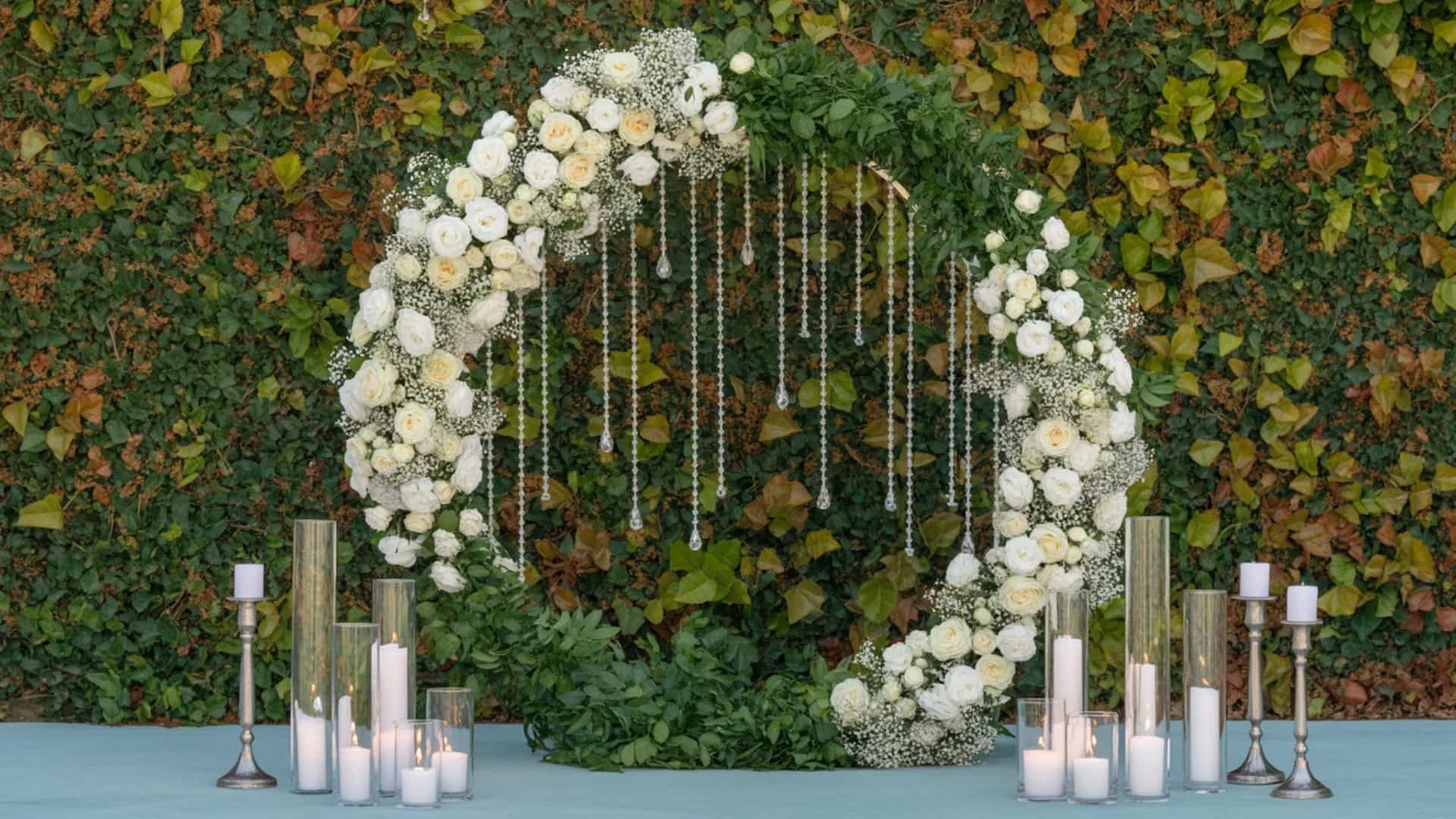 White floral ring arch with hanging crystals and candles before a leafy wall.