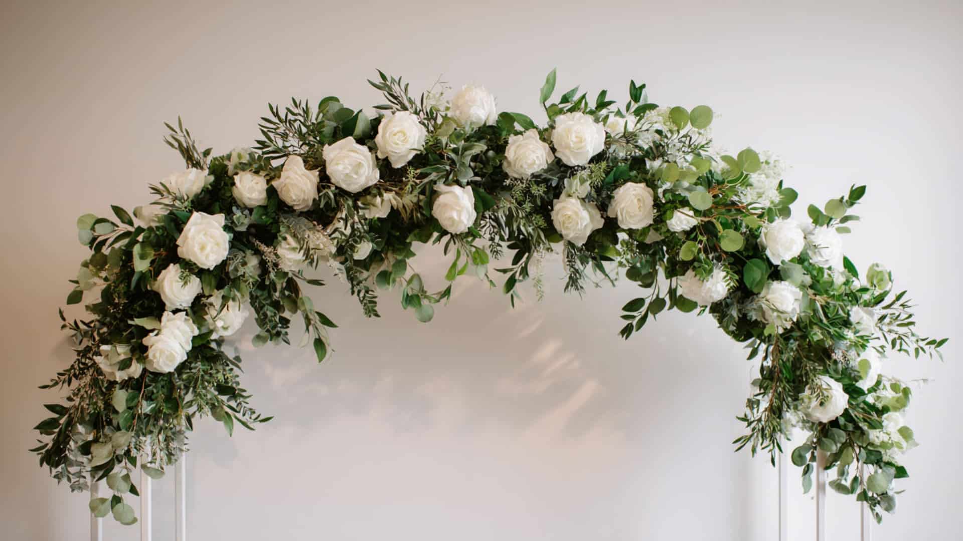 White rose and greenery wedding arch arrangement against a soft neutral wall.