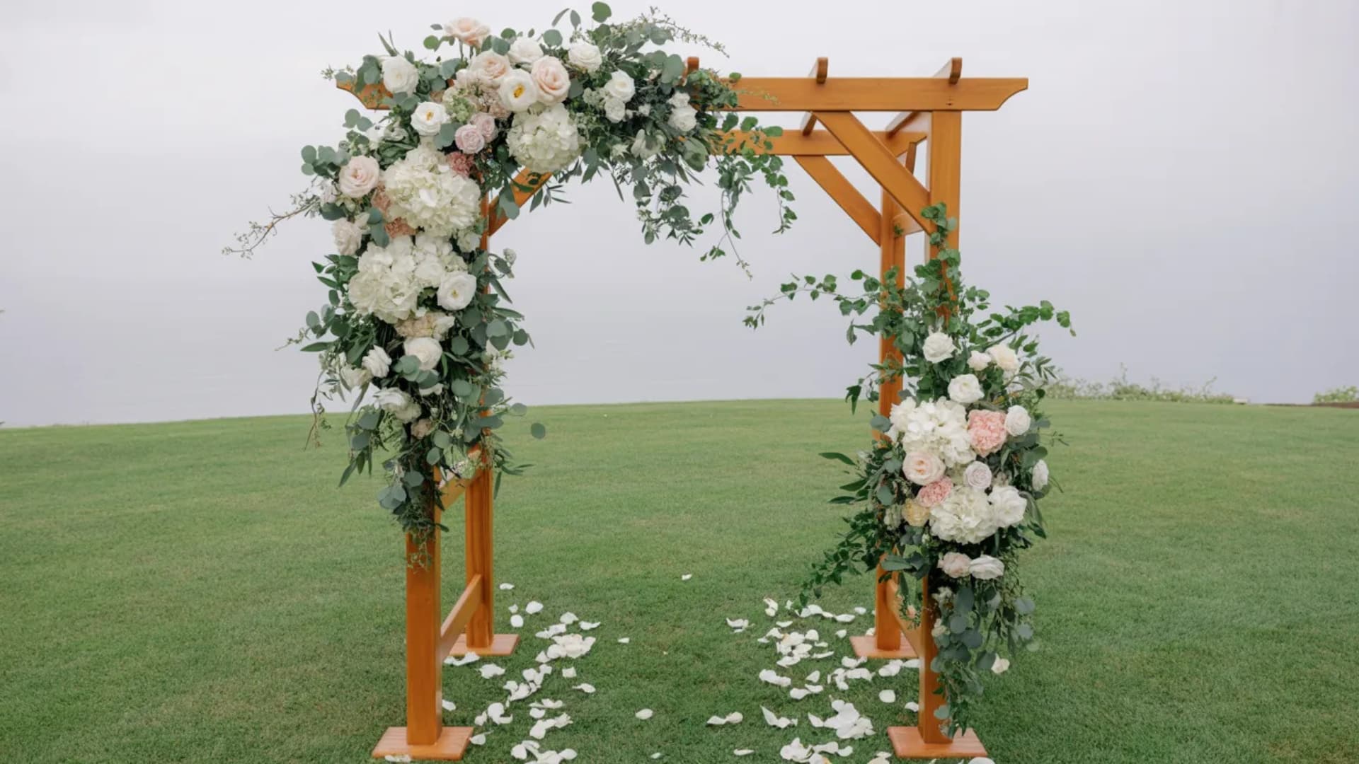 Wooden wedding arch with white flowers and greenery on a grassy cliffside lawn.