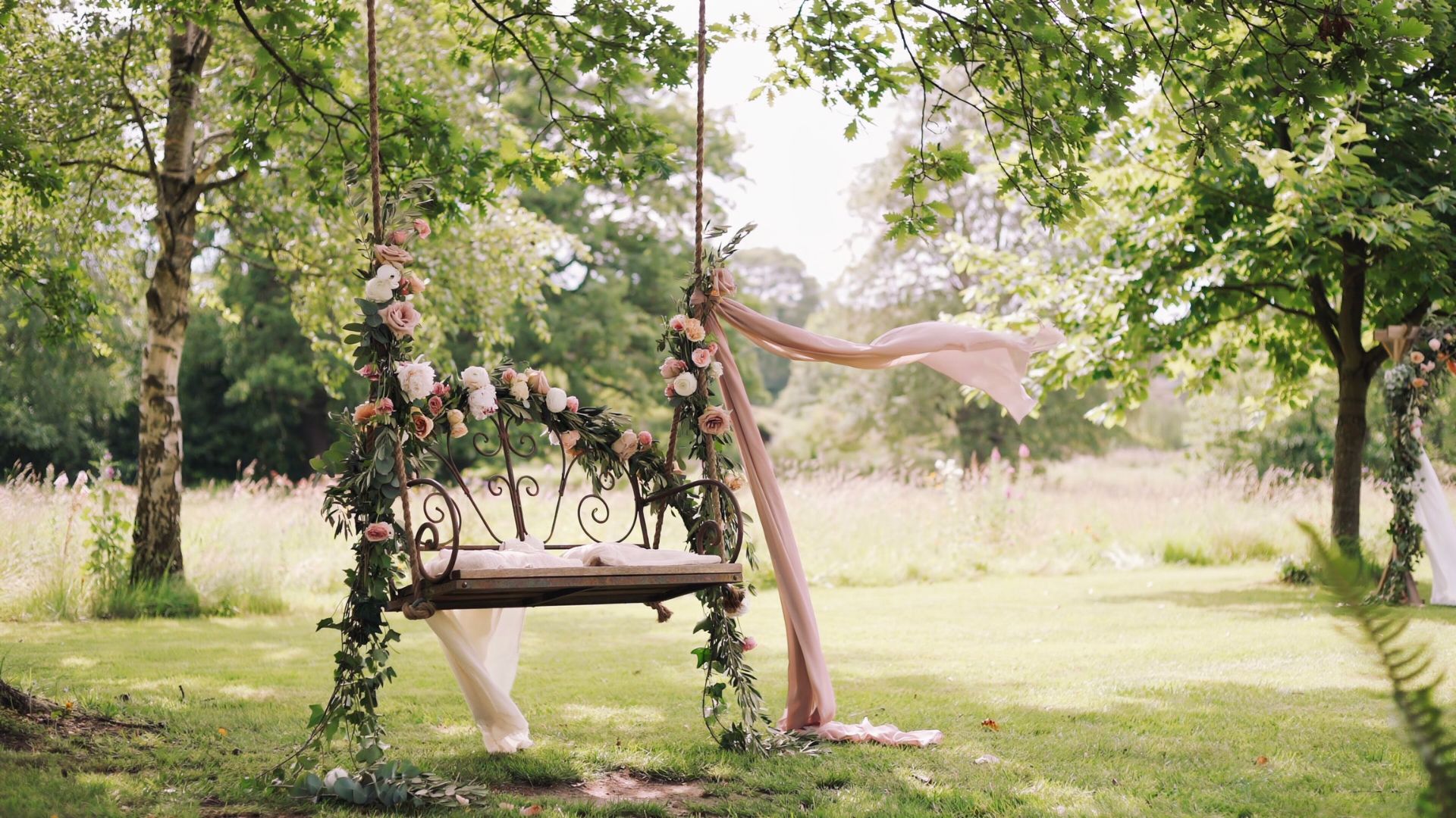 Floral garden swing with blush fabric and greenery hangs from a tree in a romantic countryside wedding setting