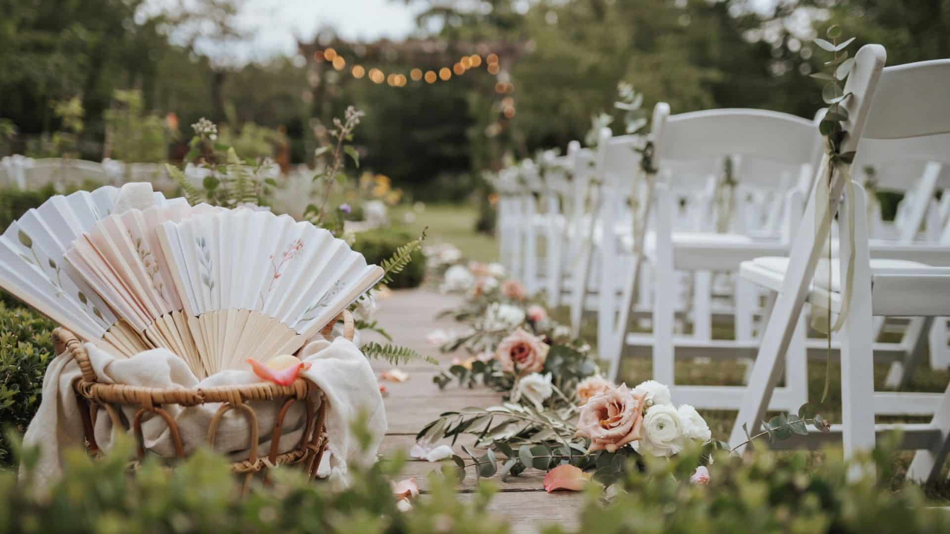 Floral hand fans rest in a wicker basket beside white ceremony chairs and a flower-lined outdoor wedding aisle