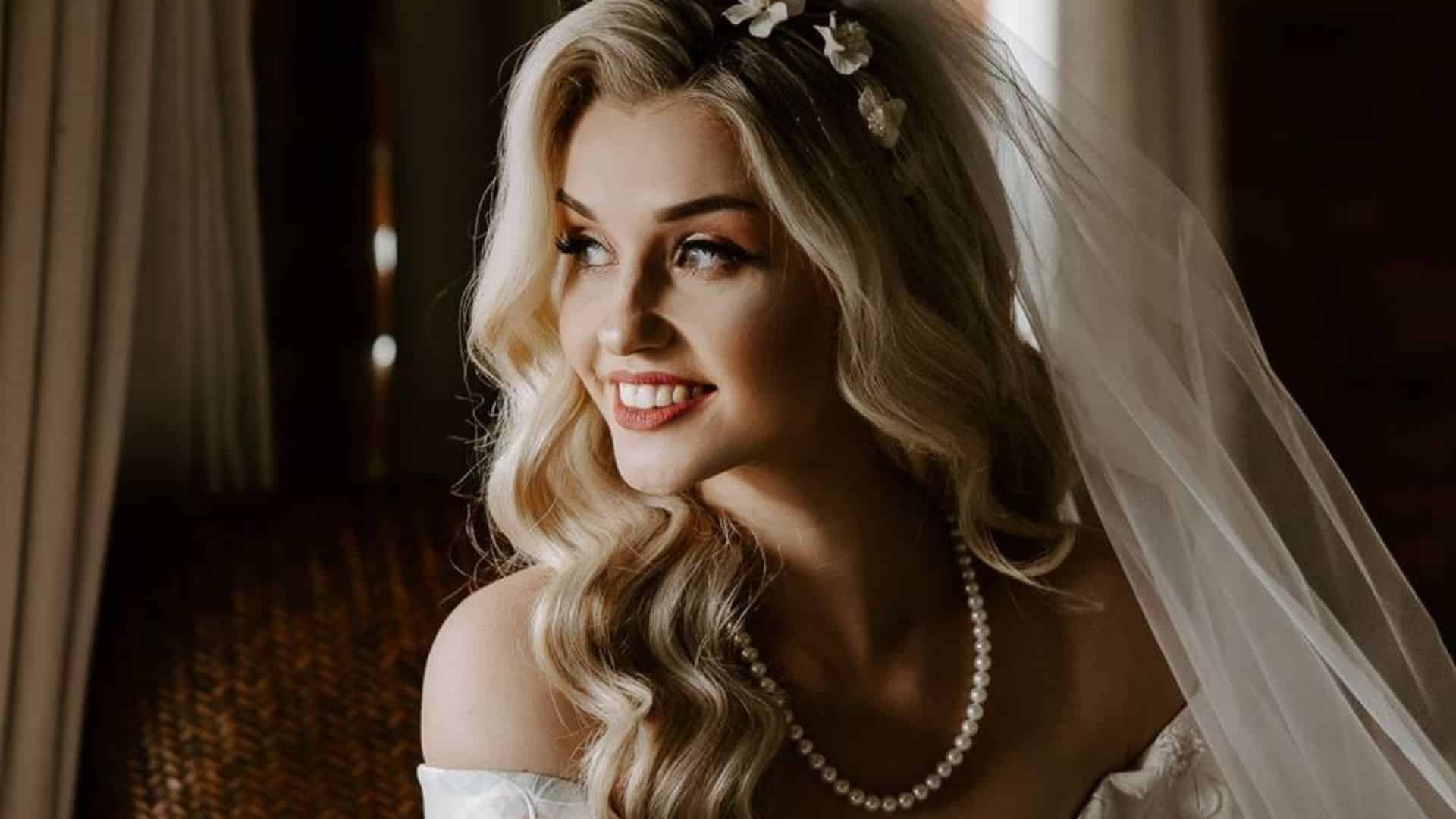 Half up half down Hollywood waves on long blonde hair with soft curls, a bridal veil, floral pins, and pearl necklace