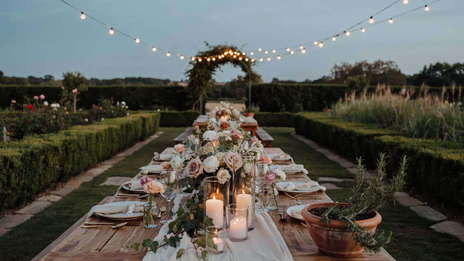 Long wooden wedding reception table with candles, pastel flowers, and string lights set in a formal garden at dusk