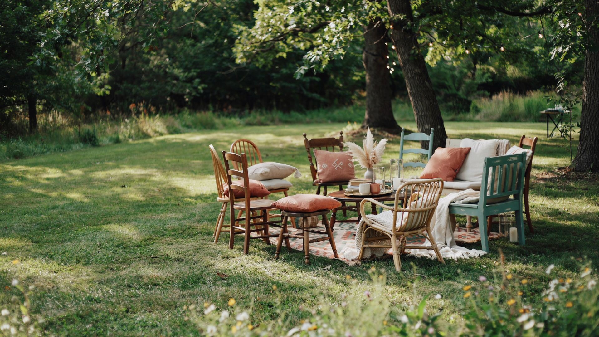 Mismatched vintage chairs, cushions, and a patterned rug create a cozy outdoor wedding lounge beneath trees