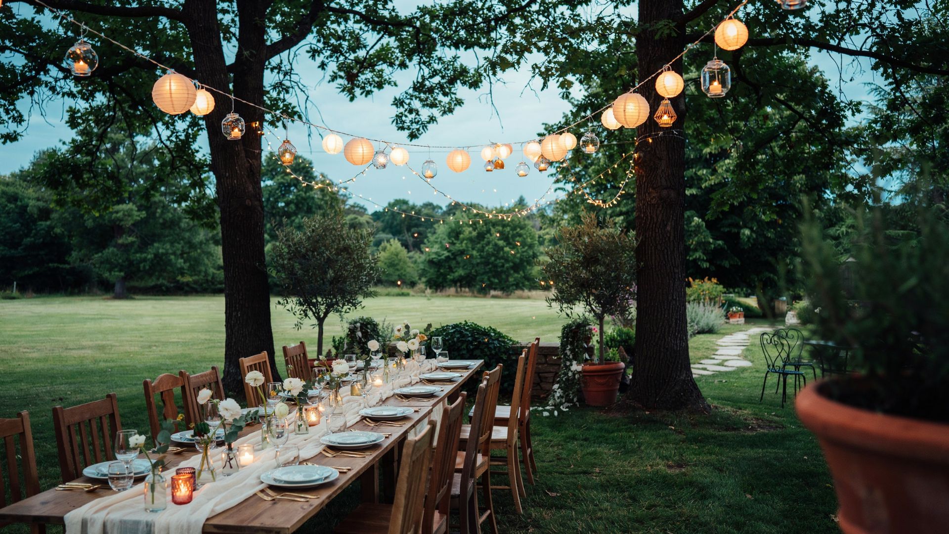 Outdoor wedding dinner table beneath glowing paper lanterns and string lights strung between large garden trees