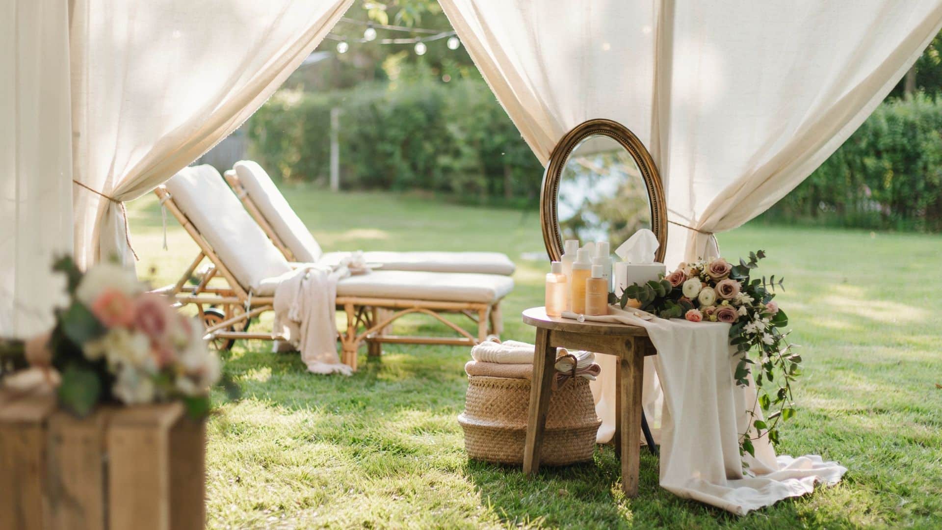 Romantic outdoor wedding lounge tent with flowing white curtains, lounge chairs, mirror, and floral decor