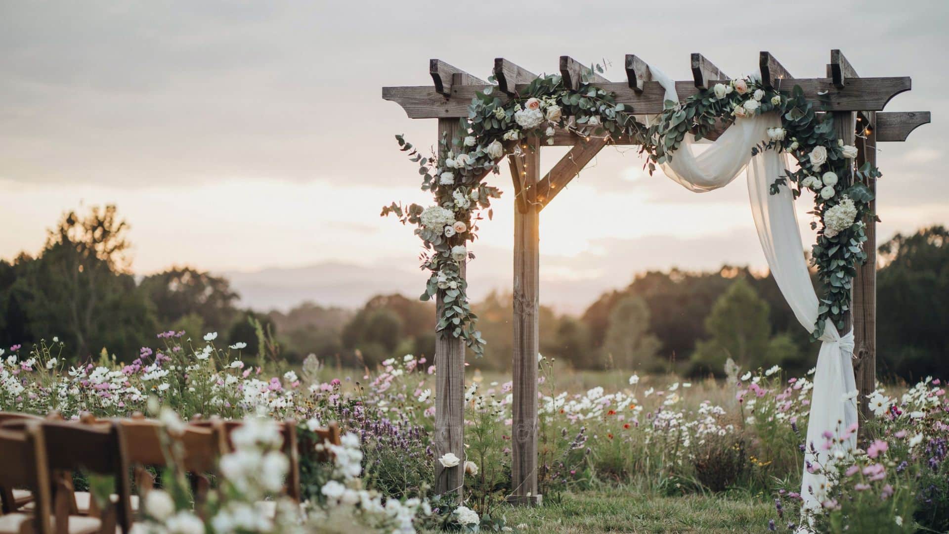 Wooden pergola draped with white fabric, greenery, and flowers overlooks a wildflower field at sunset