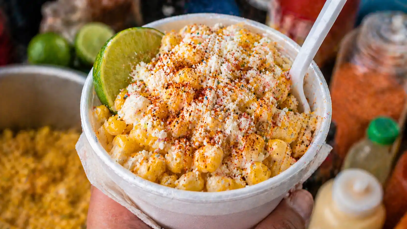 Cup of warm esquites topped with creamy sauce, crumbled cheese, chili powder, lime, and served with a spoon.