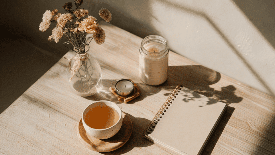 Flat lay of self-care essentials including a checklist journal, tea, candle, and flowers on a table.