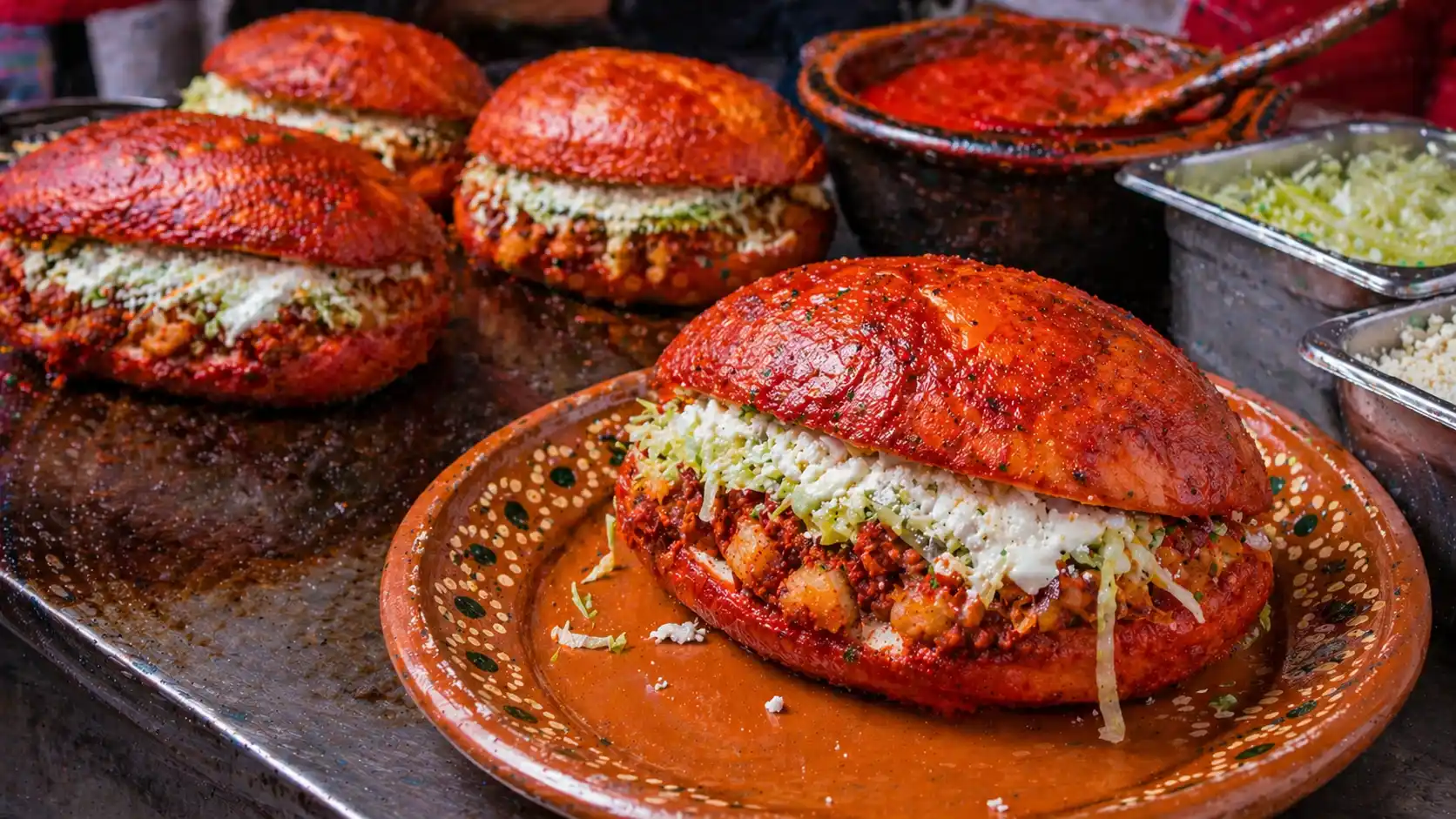 Red chili-soaked pambazo filled with potatoes and chorizo, topped with lettuce and crema beside a hot griddle.